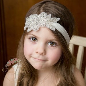 Pu&ograve; includere: Una bambina che indossa una fascia bianca con un grande motivo floreale argentato scintillante. La fascia &egrave; realizzata in un materiale morbido ed elastico, perfetta per un'occasione speciale.