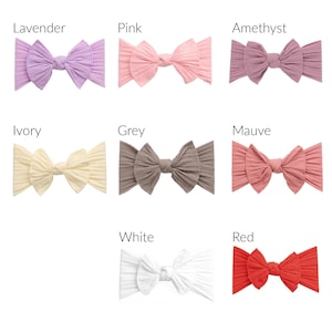 May include: A collection of eight ribbed fabric headbands in various colors, including lavender, pink, amethyst, ivory, grey, mauve, white, and red. Each headband features a large bow design.