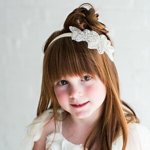 Pu&ograve; includere: Una bambina con lunghi capelli rossi indossa una fascia bianca con un motivo floreale argentato e strass. La fascia &egrave; indossata sopra la fronte ed &egrave; centrata sulla testa.