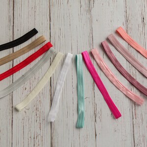 Pu&ograve; includere: Assortimento di 14 elastici per capelli colorati in varie tonalit&agrave; di rosa, rosso, beige, grigio, bianco e blu. Gli elastici per capelli sono disposti a semicerchio su una superficie in legno chiaro.