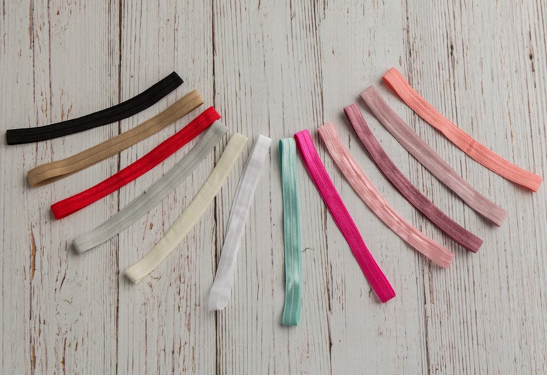 Pu&ograve; includere: Assortimento di 14 elastici per capelli colorati in varie tonalit&agrave; di rosa, rosso, beige, grigio, bianco e blu. Gli elastici per capelli sono disposti a semicerchio su una superficie in legno chiaro.