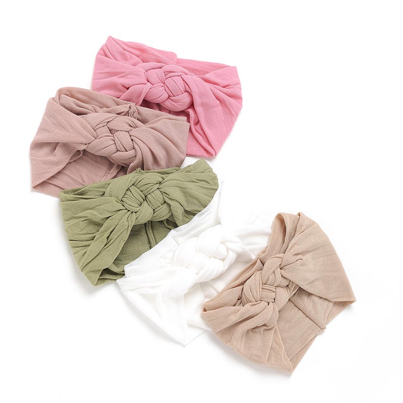 May include: A collection of knotted headbands in various colors, including pink, tan, olive green, white, and beige. The headbands are made of soft fabric and are arranged on a white background. These headbands are suitable for babies and toddlers.