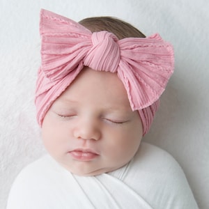 May include: A baby wearing a light pink ribbed bow headband. The headband is tied in a large bow on top of the baby's head. The baby is wrapped in a white blanket and has their eyes closed.