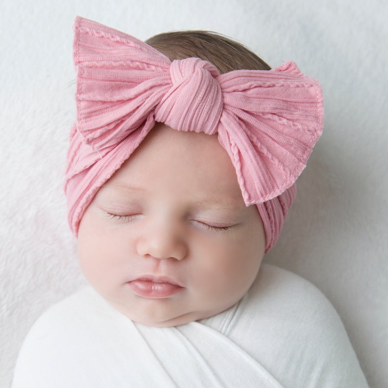 May include: A baby wearing a light pink ribbed bow headband. The headband is tied in a large bow on top of the baby's head. The baby is wrapped in a white blanket and has their eyes closed.