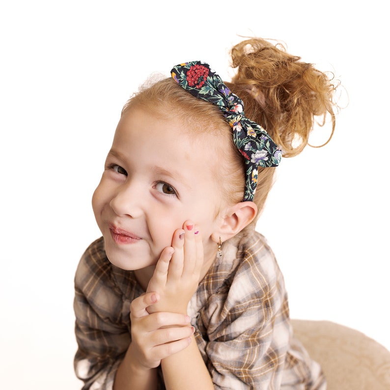 Peut inclure: Une jeune fille aux cheveux roux boucl&eacute;s portant une chemise &agrave; carreaux et un bandeau floral. Elle sourit et regarde la cam&eacute;ra.