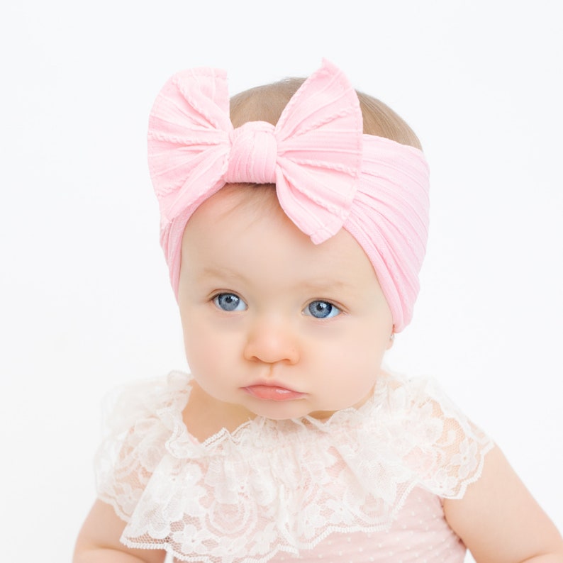 May include: A baby wearing a light pink ribbed fabric headband with a large bow. The baby has blue eyes and is wearing a pink dress with a white lace collar.