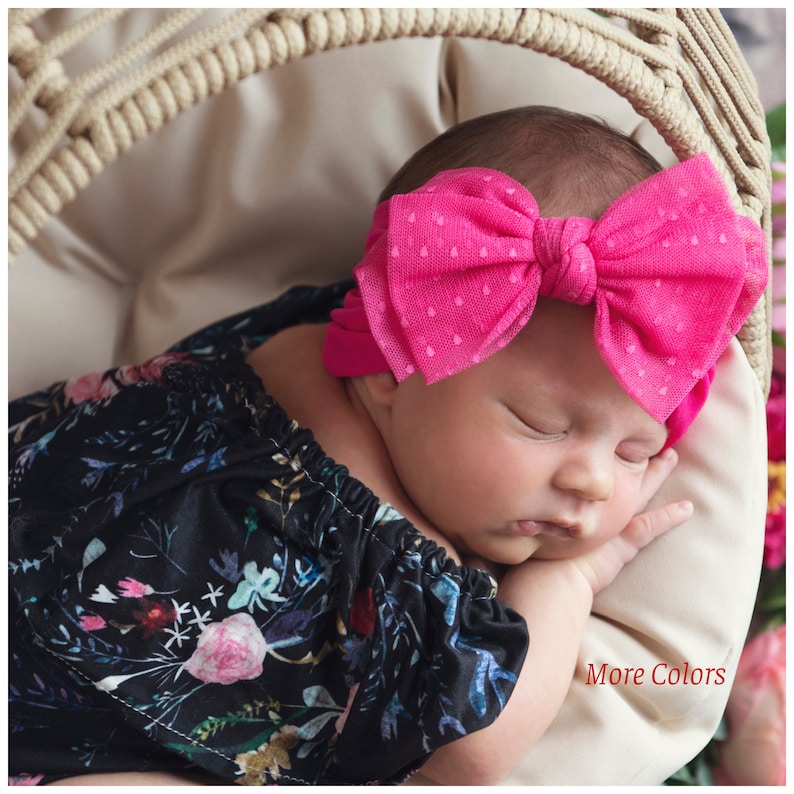 Pu&ograve; includere: Una neonata che dorme in un cesto di vimini, indossando un completo nero con stampa floreale e un cerchietto con fiocco rosa a pois. Il testo "More Colors" &egrave; visibile nell'angolo in basso a destra dell'immagine.