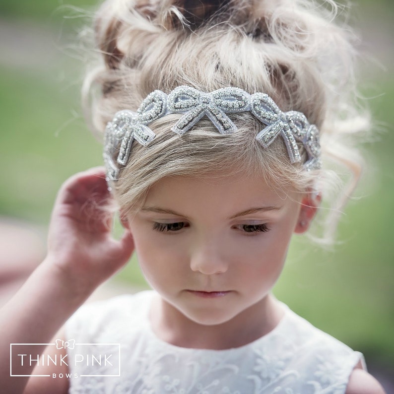 May include: A silver headband with bow detailing. The headband is adorned with small, sparkling embellishments. The image shows a young person wearing the headband, with the text "THINK PINK BOWS" in the lower left corner.