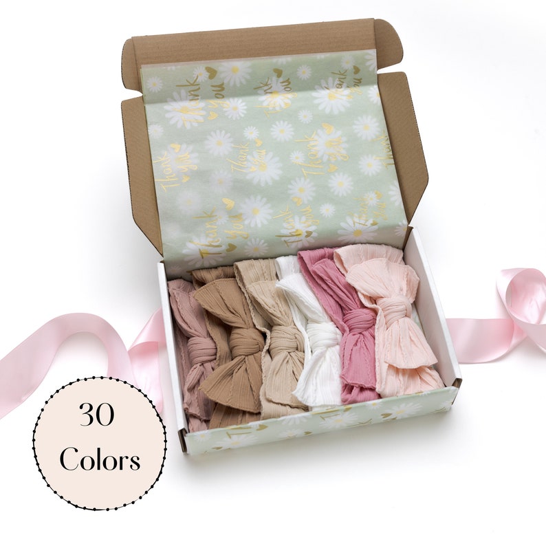 May include: An open cardboard box filled with assorted fabric bows in shades of pink, beige, and white. The box is lined with tissue paper featuring a daisy pattern and gold text. A circular graphic reads "30 Colors".