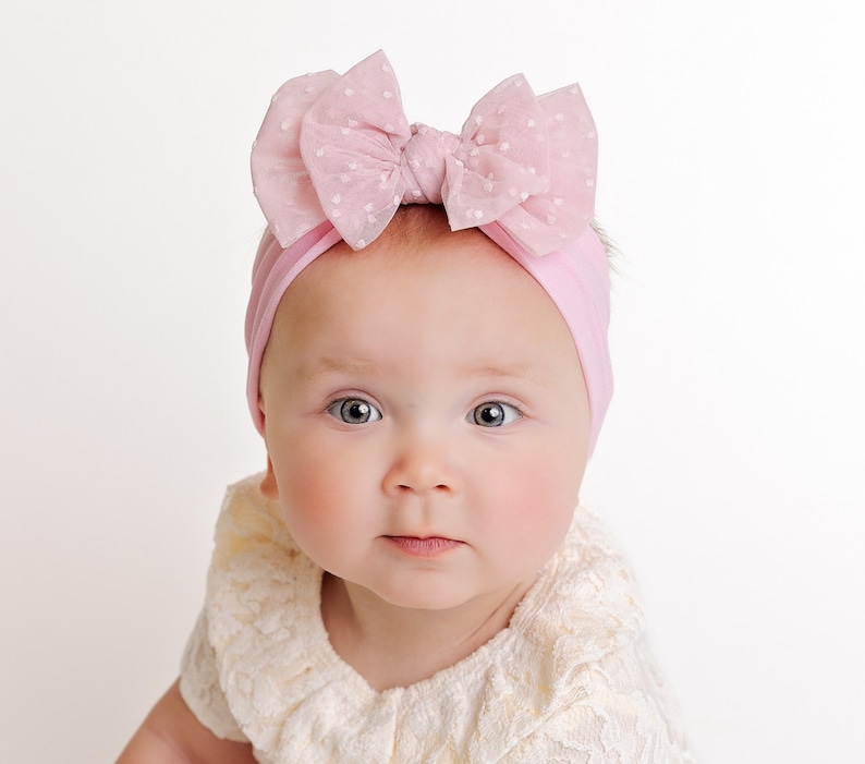 Pu&ograve; includere: Un bambino che indossa una fascia rosa con un grande fiocco rosa. La fascia &egrave; realizzata in un materiale morbido ed elastico e il fiocco &egrave; realizzato in un tessuto delicato e trasparente. Il bambino sta guardando la telecamera con un'espressione seria.