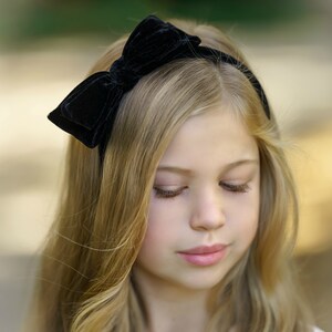 May include: A young girl with long blonde hair wearing a black velvet headband with a bow.