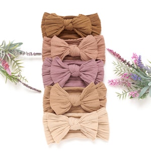 May include: Five soft, textured headbands in varying shades of brown, pink, and beige are arranged vertically. Each headband features a large, decorative bow. The headbands are displayed on a white background, with sprigs of lavender and greenery on either side.