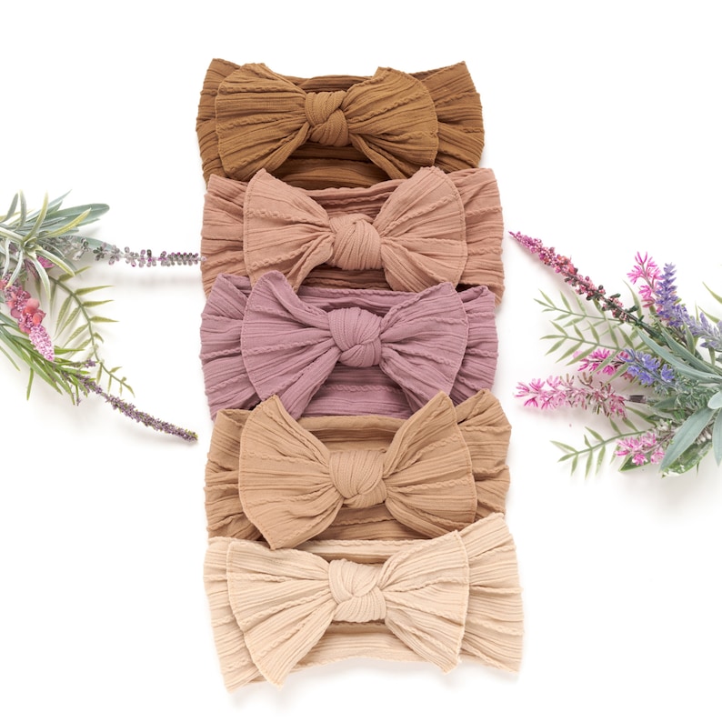 May include: Five soft, textured headbands in varying shades of brown, pink, and beige are arranged vertically. Each headband features a large, decorative bow. The headbands are displayed on a white background, with sprigs of lavender and greenery on either side.