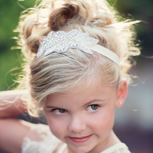 Pu&ograve; includere: Una bambina con i capelli biondi che indossa una fascia bianca con un ornamento argentato e strass. La fascia &egrave; indossata sopra la fronte e i suoi capelli sono raccolti in uno chignon disordinato.
