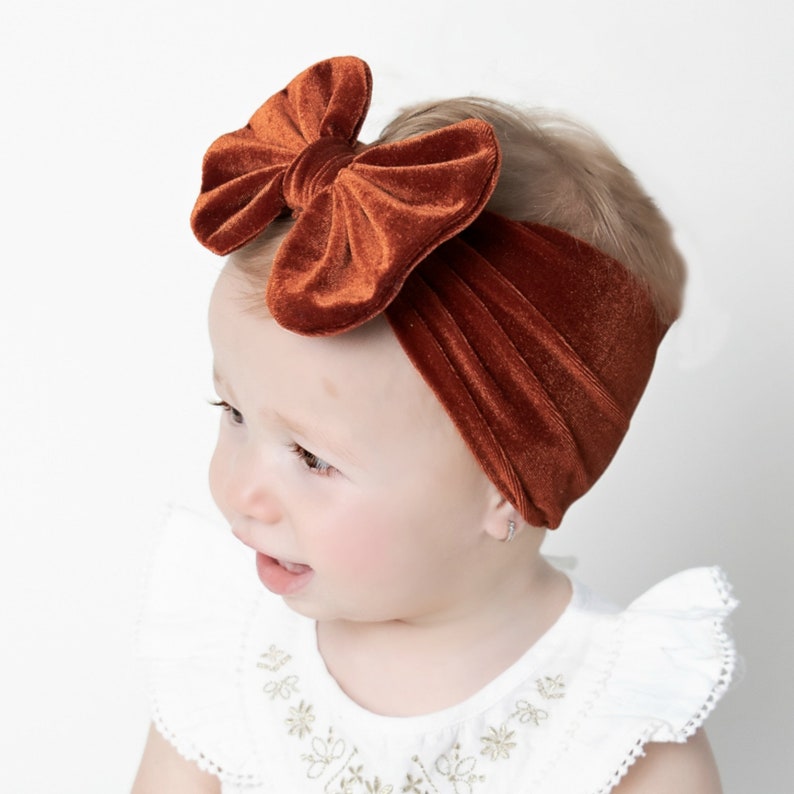 May include: A brown velvet headband with a large bow on top. The headband is worn by a baby.