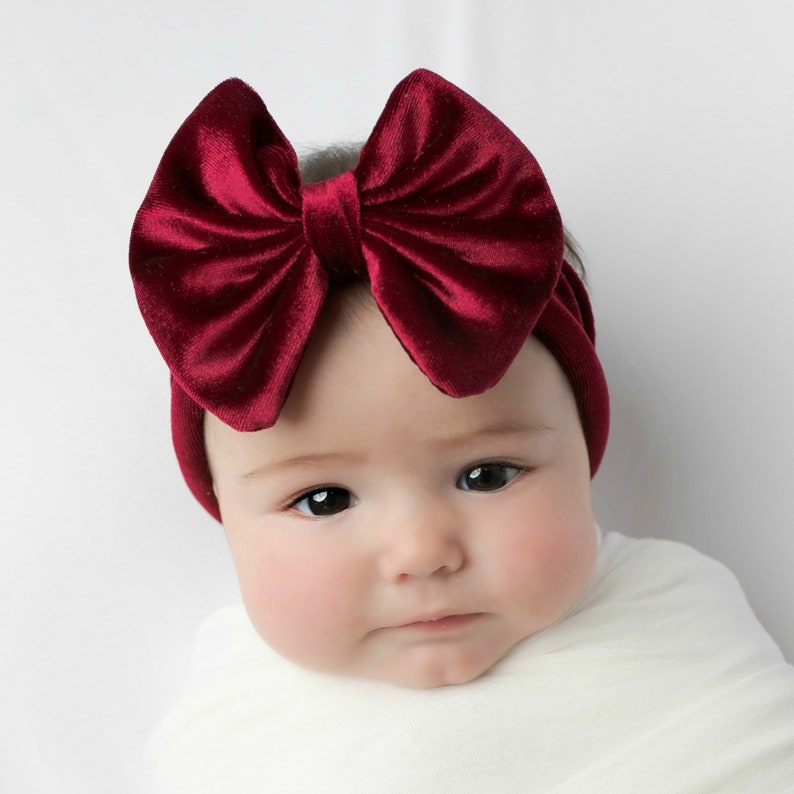 May include: A baby wearing a burgundy velvet headband with a large bow. The headband is made of soft, stretchy material and is perfect for newborns and infants.