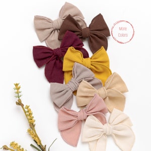 May include: A set of ten fabric hair bows in various colors, including beige, brown, burgundy, mustard yellow, and pink. The bows are arranged in a semi-circle on a white background. The text "More Colors" is written in red on a white circle in the upper right corner of the image.