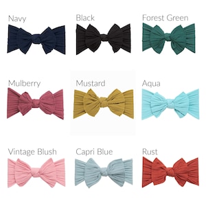May include: A collection of nine ribbed fabric headbands with knotted bows, displayed in a grid. Colors include navy, black, forest green, mulberry, mustard, aqua, vintage blush, capri blue, and rust, each labeled with its respective color name.