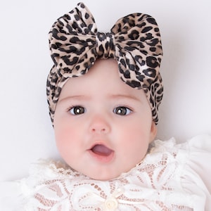 May include: A baby wearing a white lace dress with a brown and black leopard print headband with a large bow.