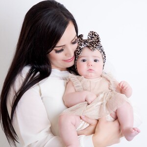May include: A woman with long dark hair holds a baby girl wearing a beige dress and a leopard print headband. The baby is looking at the camera.