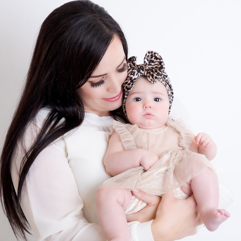 May include: A woman with long dark hair holds a baby girl wearing a beige dress and a leopard print headband. The baby is looking at the camera.
