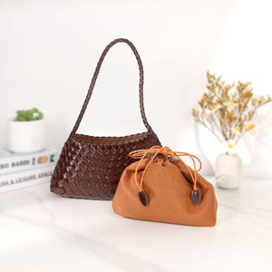 May include: A brown woven leather handbag with a braided handle stands next to a small, tan drawstring pouch. The handbag has a trapezoid shape. The pouch has a drawstring closure with brown leather accents.