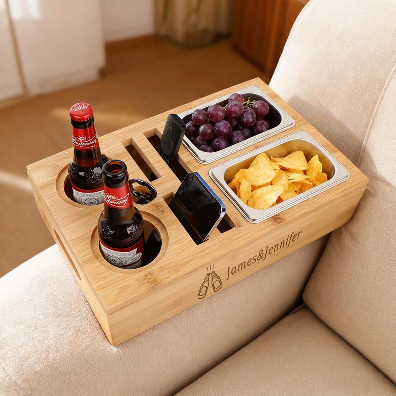 May include: A bamboo sofa arm tray with two beer bottles, two stainless steel food containers, and slots for a phone and remote. The tray holds grapes and chips. The text "James & Jennifer" is engraved on the front.