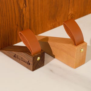 May include: Two wooden doorstops with brown leather handles. One doorstop is a dark wood with the text "Merry Christmas" engraved on the side. The other doorstop is a light wood. Both have a gold-colored metal accent.