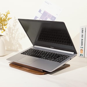 May include: A silver Acer laptop is open on a wooden laptop stand. The stand is angled, and the laptop screen is tilted upwards. A vase with yellow flowers and a book are in the background.