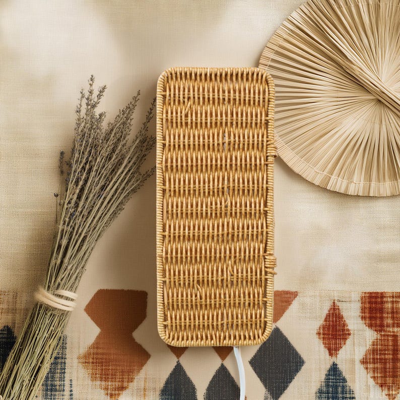 Pu&ograve; includere: Un cestino rettangolare in rattan intrecciato con un cavo bianco, un mazzo di lavanda secca e un ventaglio decorativo. Il cestino &egrave; marrone chiaro e ha una superficie strutturata. Il ventaglio &egrave; fatto di fibre naturali. Gli oggetti sono su un tessuto fantasia.