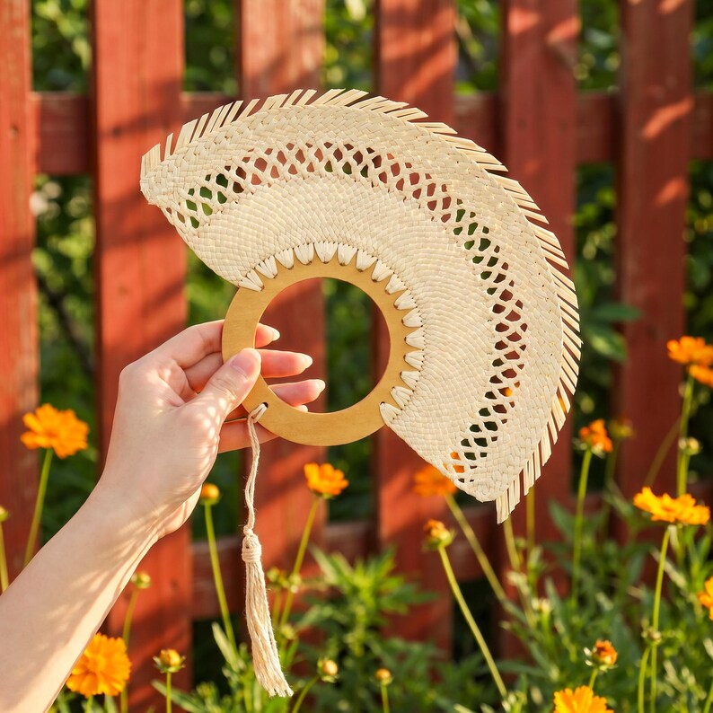 Puede incluir: Un abanico de mano hecho de fibras naturales tejidas. El abanico tiene un mango circular de madera y una borla decorativa. El dise&ntilde;o del abanico incluye patrones calados y un borde festoneado. El fondo presenta flores naranjas y una valla roja.