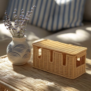 Pu&ograve; includere: Un contenitore rettangolare in vimini chiaro con coperchio &egrave; appoggiato su un tavolo intrecciato. Un piccolo vaso bianco con disegni di onde blu contiene rametti di lavanda. Un cuscino a righe blu e bianco sullo sfondo.