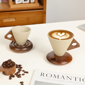May include: Two beige ceramic coffee pour-over cones with wooden handles and bases. One cone is filled with coffee and latte art. A wooden scoop of coffee beans and scattered beans are in the foreground.