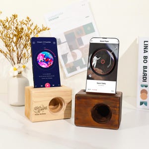 May include: Two wooden phone holders, one light and one dark, each with a circular speaker hole. The light wood holder has "Happy Birthday" text. Phones display music apps with colorful graphics. Decorative items are in the background.