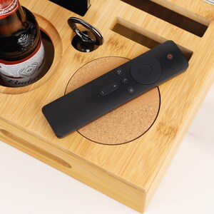 May include: A bamboo serving tray with a black remote control, a bottle of beer, and a metal ring holder. The remote is on a cork coaster. The tray has cutouts for items.