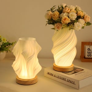 Puede incluir: Dos lámparas decorativas con un brillo cálido. Una lámpara tiene una pantalla de vidrio esmerilado ondulado, y la otra es una lámpara en forma de jarrón con un diseño en espiral. Ambas lámparas están sobre bases de madera. Un ramo de rosas está en la lámpara en forma de jarrón.