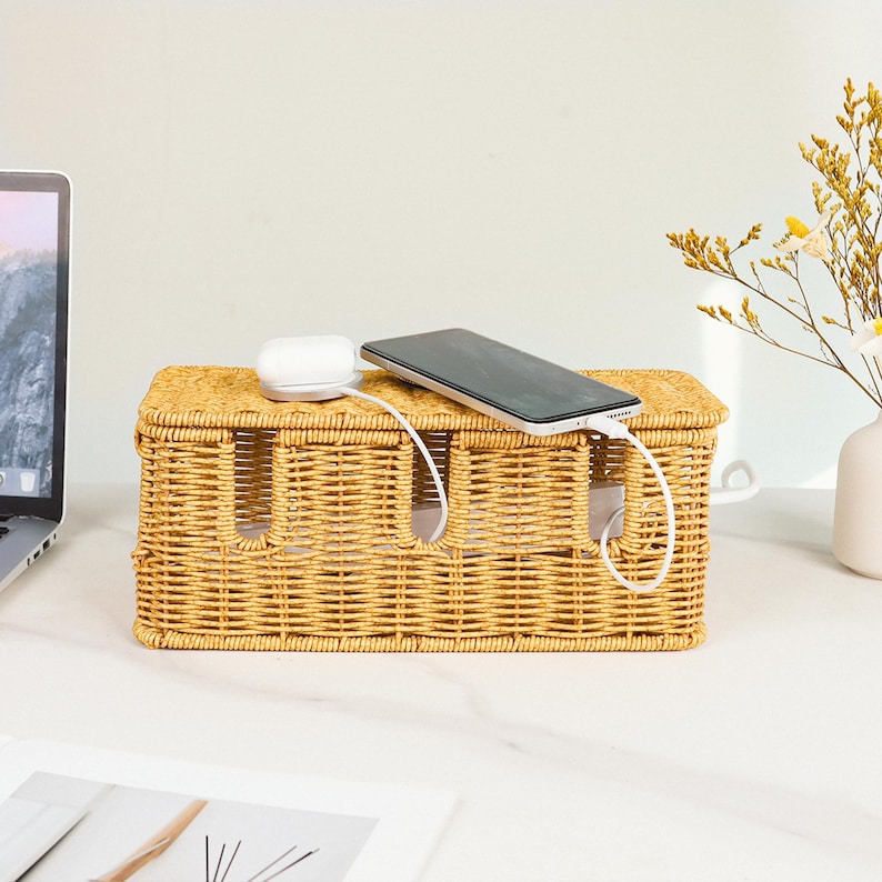 Pu&ograve; includere: Un organizer per cavi in vimini intrecciato di colore marrone chiaro. La scatola rettangolare ha tre fessure per cavi e un coperchio. Uno smartphone e auricolari wireless sono appoggiati sopra, collegati all'organizer.