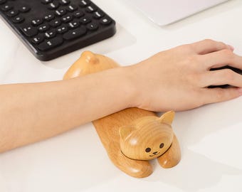 May include: A wooden cat-shaped wrist rest for a computer mouse. The wrist rest is light brown with a smooth finish and features a carved cat face with small black eyes and a smiling mouth. The cat is lying down, supporting the wrist.