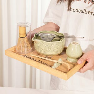 Könnte beinhalten: Ein Matcha-Teeset auf einem Bambustablett. Das Set beinhaltet eine grüne Keramikschale mit floralen Mustern, einen Schneebesen, einen Schneebesenhalter, ein Sieb und einen Bambuslöffel. Auf dem Tablett steht "Matcha Moment".