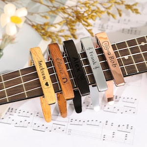 Op de afbeelding: Een verzameling van zes ukelele capo's in verschillende kleuren, waaronder houtnerf, zwart en zilver, elk gegraveerd met een naam. De capo's zijn bevestigd aan een ukelele toets, rustend op bladmuziek.