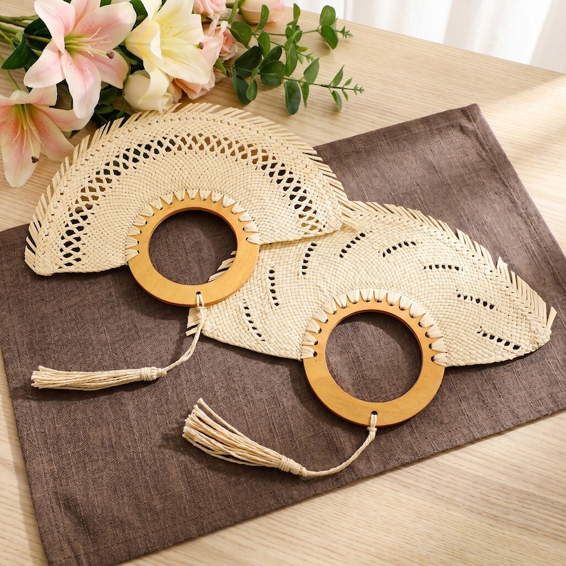 Puede incluir: Dos abanicos de mano de paja tejida con mangos de madera y borlas. Los abanicos son de color beige claro con forma semicircular y cortes decorativos. Los mangos son redondos y de madera clara. Los abanicos est&aacute;n sobre una superficie marr&oacute;n con flores rosas en el fondo.