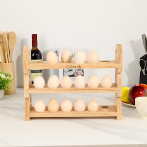 May include: A three-tiered wooden egg rack holding 12 eggs. The rack is made of light-coloured wood and has a rectangular shape. The eggs are light brown. The brand name "Frank" is printed on the middle tier. A bottle of wine and utensils are in the background.