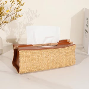 May include: A rectangular tissue box with a woven straw body and brown leather trim. The top features a wooden handle with the name "Frank" engraved. White tissues are visible.