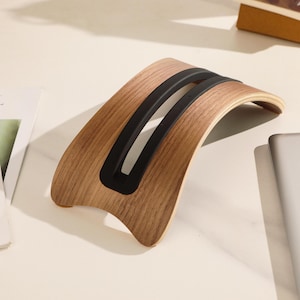 Pode incluir: Suporte de madeira para laptop com design curvo. O suporte é feito de madeira marrom clara com duas tiras de borracha preta na parte superior. O suporte foi projetado para elevar um laptop para melhor ergonomia.