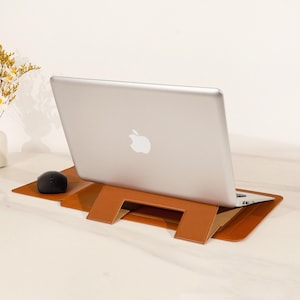 May include: A silver laptop with the Apple logo is propped up on a brown leather laptop stand. A black computer mouse sits to the left of the laptop. The stand is on a light-colored surface.