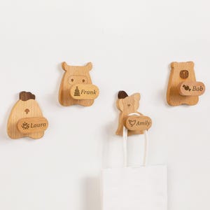 Könnte beinhalten: Vier hölzerne Wandhaken in Tierform, darunter ein Nilpferd und ein Bär. Jeder Haken trägt einen Namen, der auf einer kleinen Holzplakette eingraviert ist: Laura, Frank, Amily und Bob. An einem Haken hängt eine weiße Papiertüte.
