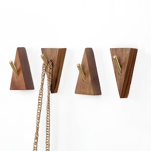 May include: Four triangular wooden wall hooks with brass-coloured metal hooks. One hook holds a gold-coloured chain. The hooks are made of dark brown wood and are mounted on a white wall.