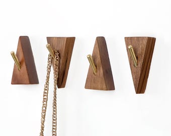 May include: Four wooden wall hooks in a triangular shape with brass-colored metal hooks. One hook holds a gold-colored chain. The hooks are made of dark brown wood and are mounted on a white wall.