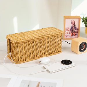 Pu&ograve; includere: Un contenitore per cavi in vimini intrecciato di colore marrone naturale. Uno smartphone bianco &egrave; collegato a un pad di ricarica bianco. Un piccolo altoparlante in legno e una foto incorniciata sono anche sulla superficie bianca.