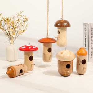 Pode incluir: Uma coleção de casas de pássaros de madeira em várias formas, incluindo cogumelos e bolotas. As casas de pássaros são pintadas em tons de madeira natural, vermelho e laranja, com pequenos orifícios de entrada e cordas para pendurar. Algumas estão penduradas, outras estão em uma superfície.